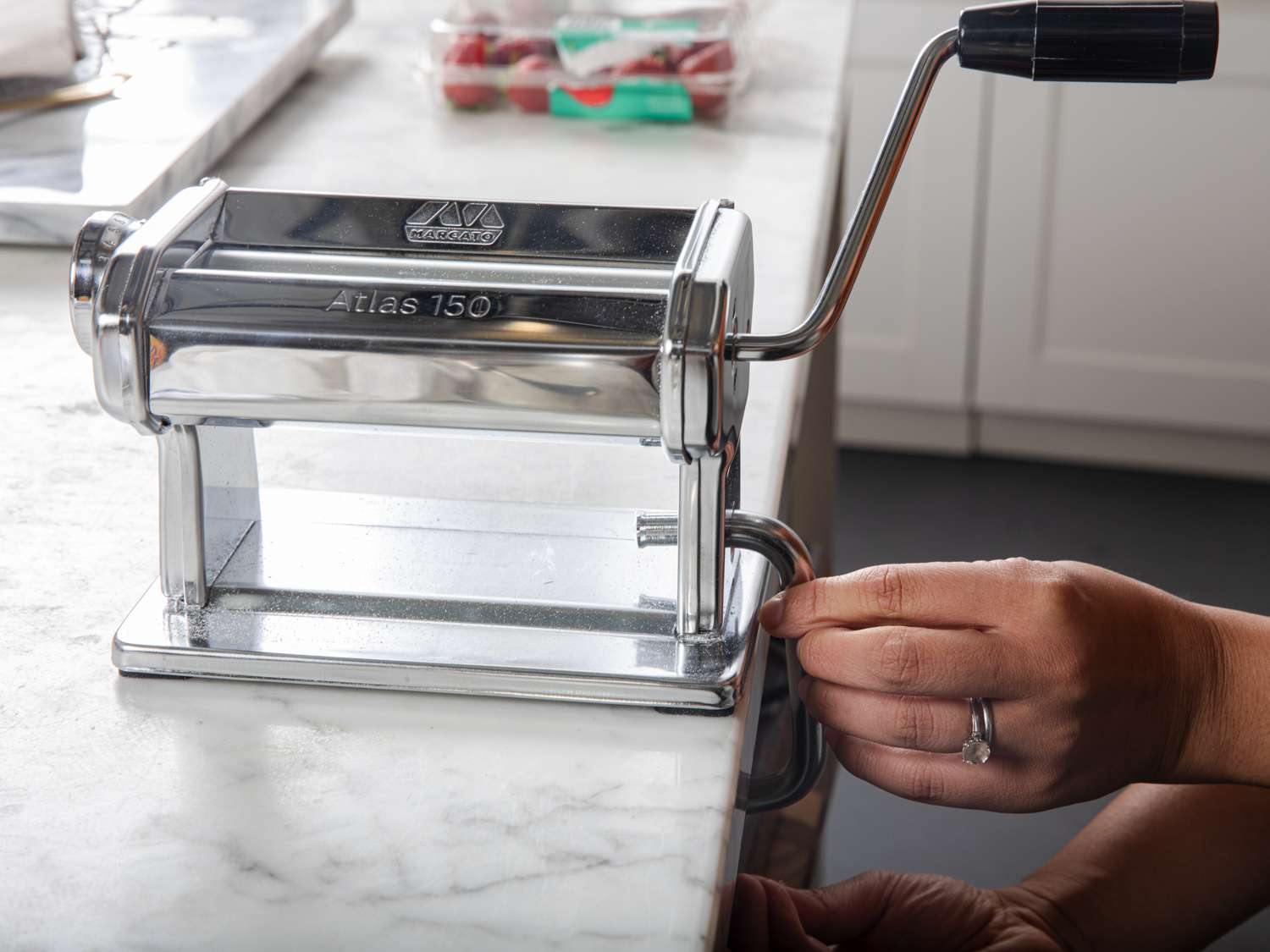 Two hands adjusting the clamp of a manual pasta maker on the side of a countertop