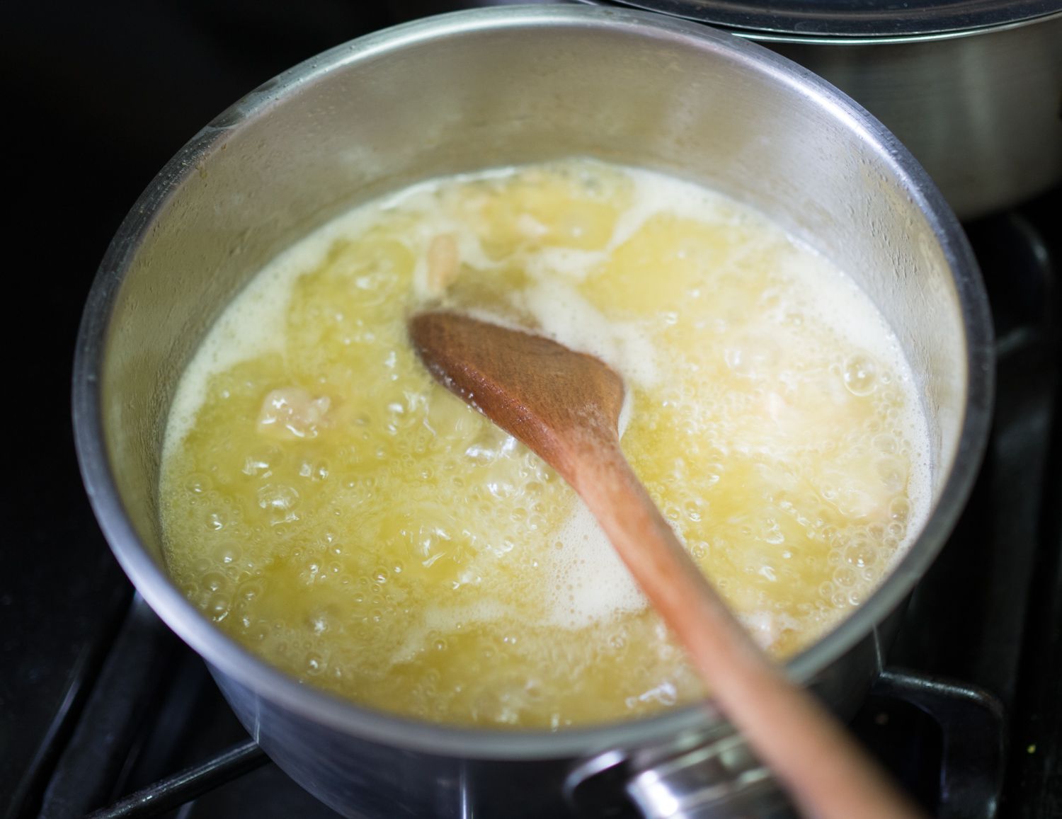 A bubbling saucepan of chicken fat being rendered on a stove. A wooden spoon is stirring the pan.