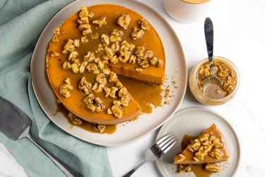 A deep orange sweet potato cheesecake drizzled with a flood of walnuts and caramel sits on a large plate with one slice missing; that slice is on a serving plate next to it.