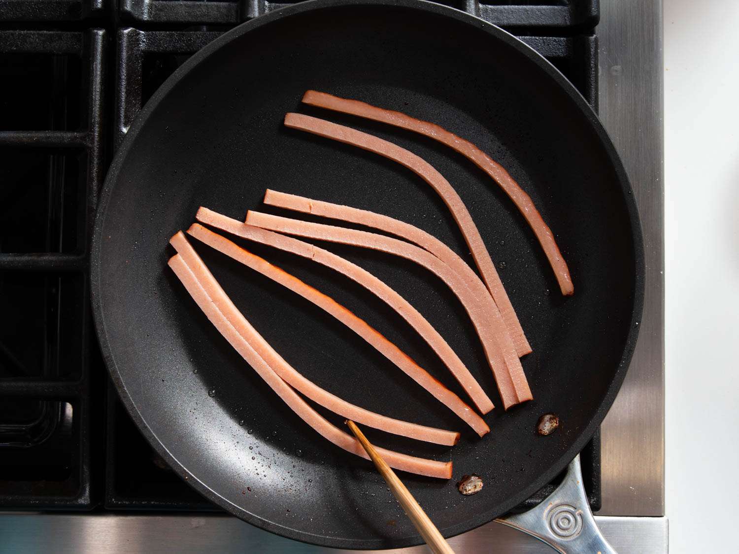 Overhead shot of thin, square strips of ham frying in a nonstick skillet.