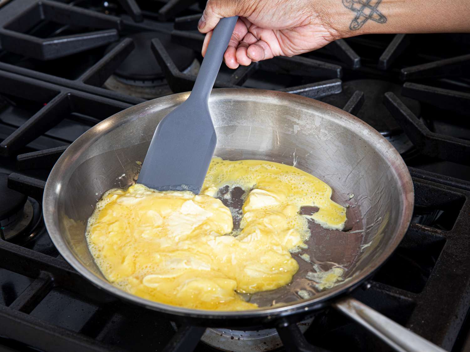 A hand holding a grey silicone spatula and scrambling eggs in a stainless steel skillet