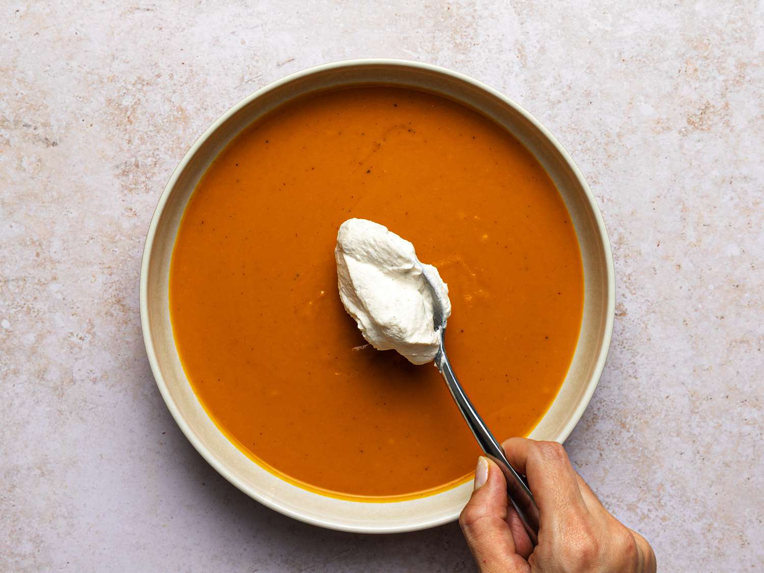 A hand spooning spiced whipped cream onto bowl of soup.