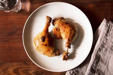 Two confit duck legs, cooked sous vide and then seared, on a white plate set on a wooden tabletop with a gray napkin and glass of water on the side.