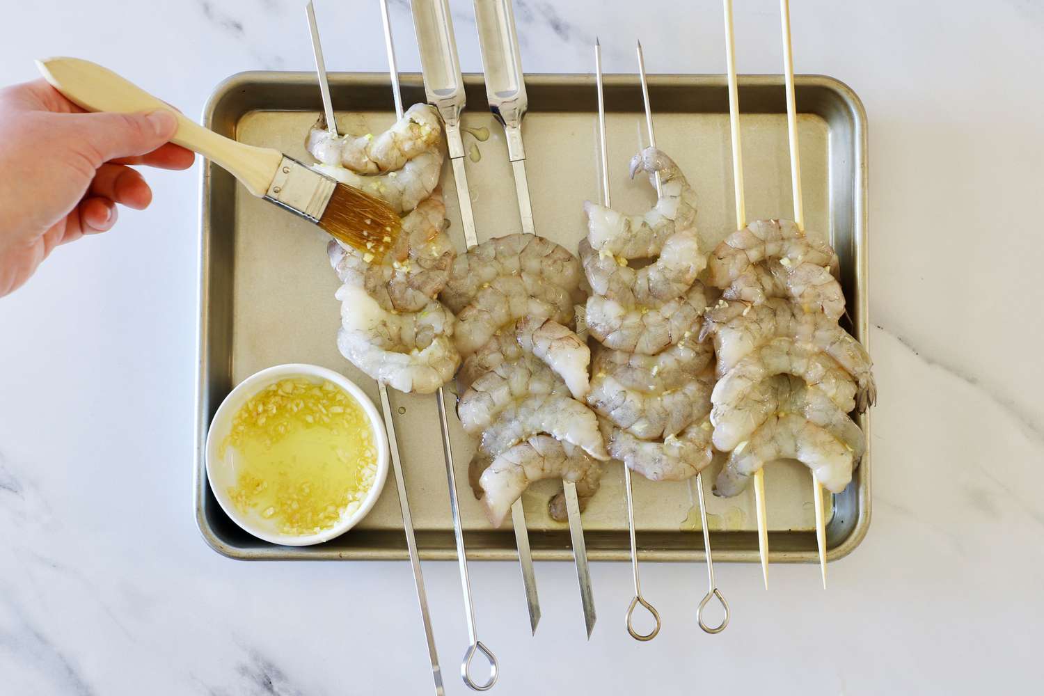 a person using a brush to baste shrimp skewers on a sheet tray