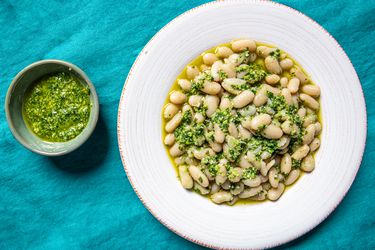 顶视图的卡尔ic scape pesto in a small bowl next to a wide rimmed, shallow bowl of cooked white beans and pesto