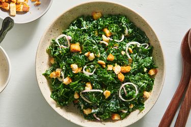 Kale caesar salad in a speckled ceramic bowl. On the right periphery are a pair of wooden serving spoons, and on the left periphery is a small bowl holding croutons.