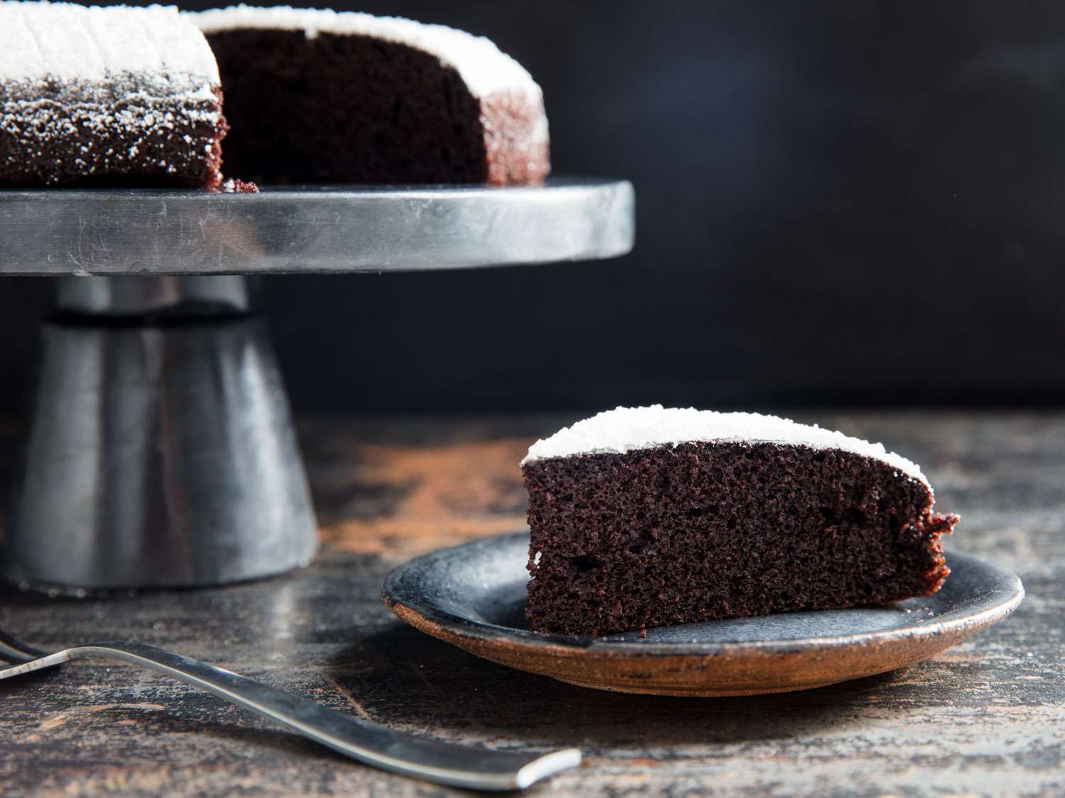 一块巧克力橄榄油蛋糕，上面撒上糖粉，背景是完整的蛋糕