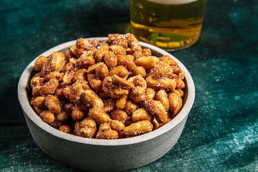 Bowl of honey roasted peanuts on a textured green fabric next to a glass of beer