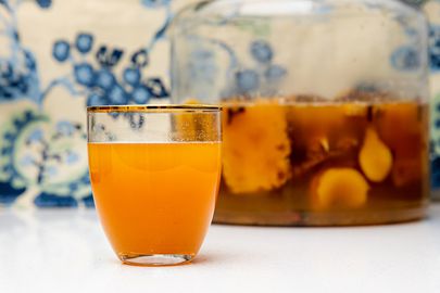 Side view of a glass of tepache next to a large batch in a gallon containter