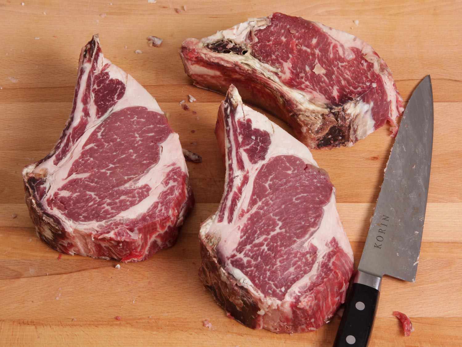 Three thick steaks on a wooden surface next to a large knife