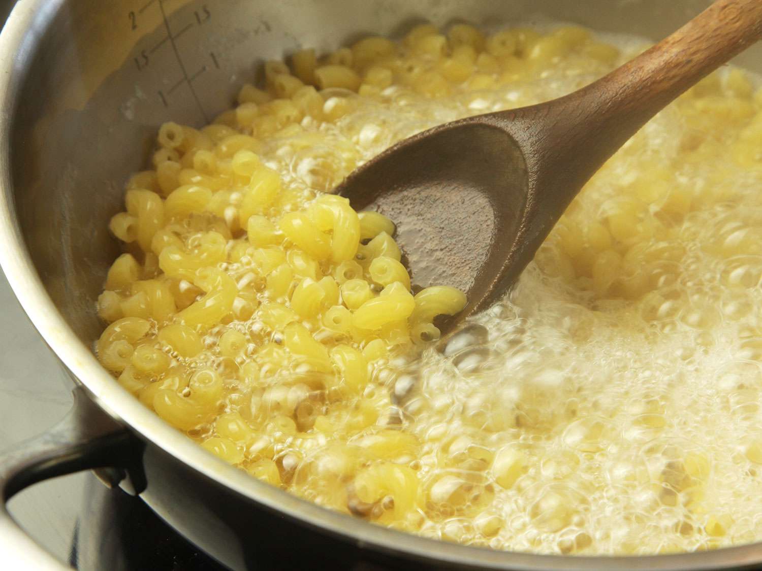 Macaroni in a boiling pot of water, being stirred. There is only just enough water to cover the pasta, which helps concentrate starch.