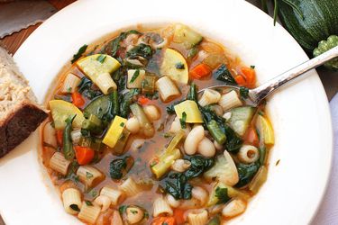 Overhead of a minestrone soup in a white bowl with a slice of bread off to the side