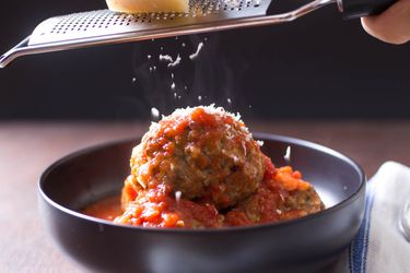 Grating Parmesan cheese on top of Italian-American meatballs in red sauce in a dark bowl.