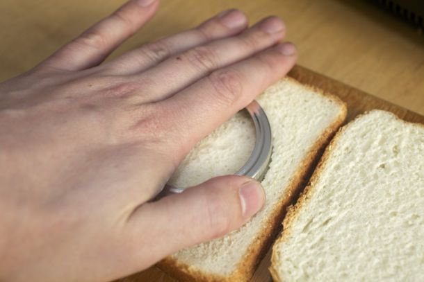 一只手用玻璃瓶上的戒指在一片三明治面包中间戳出一个洞