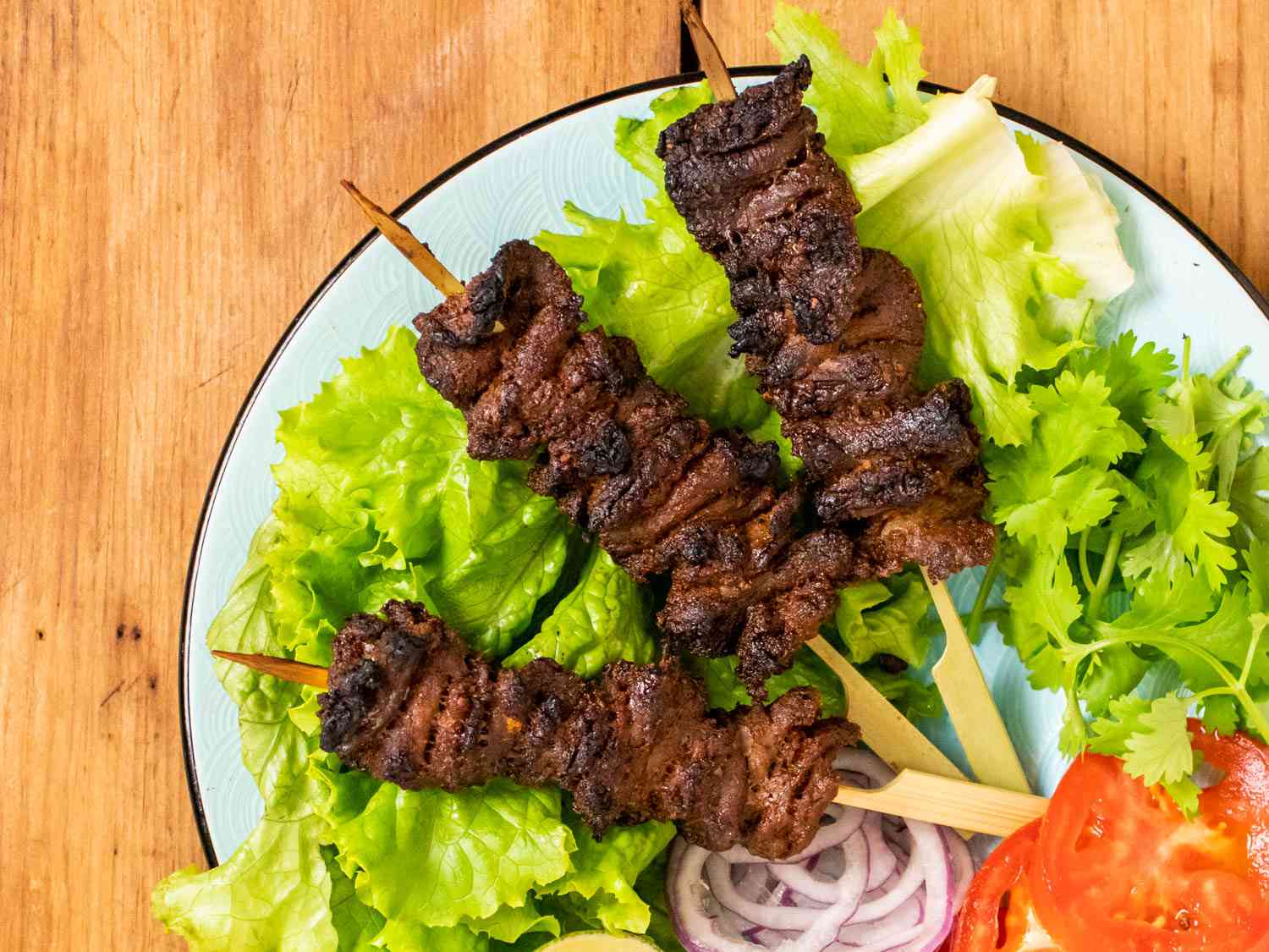 A plate with three Nigerian beef suya (spiced grilled skewers) on a bed of lettuce with sliced red onions, tomato sliced, and cilantro.