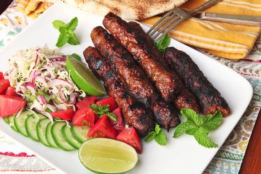 A platter of Pakistani Seekh kebabs with slaw, cucumbers tomatoes, and lime wedges.