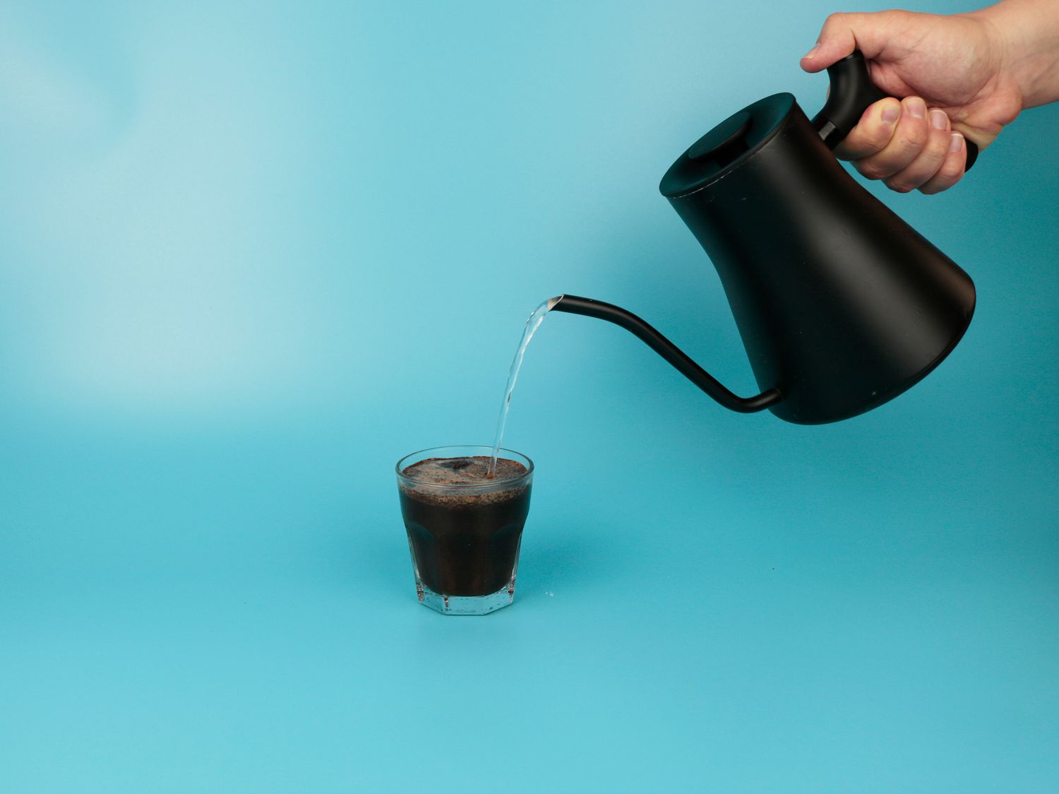 a gooseneck kettle pouring boiling water into coffee inside a glass