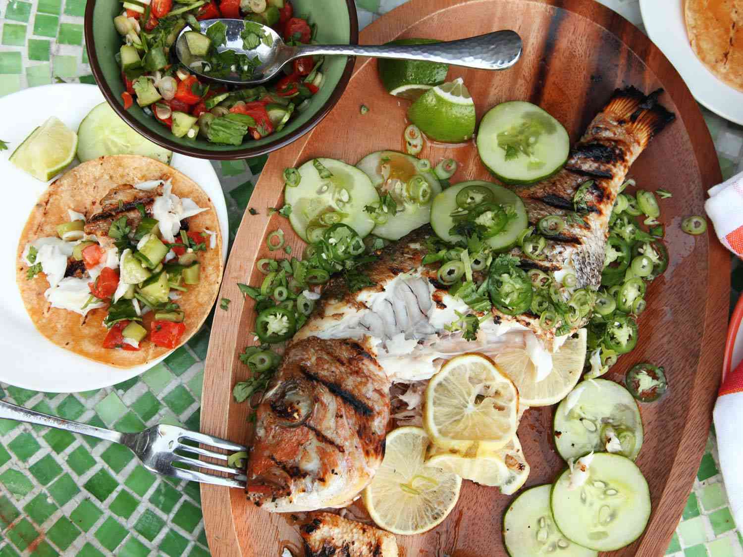烤整条鱼，配上辣椒、黄瓜片和柠檬片，旁边的盘子里有鱼肉卷和配套的食物