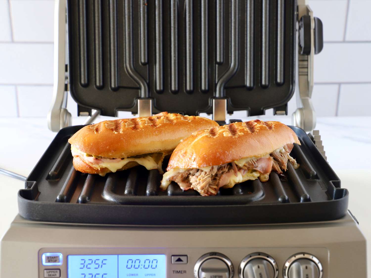 two cubano pressed sandwiches on a panini press with its lid upright
