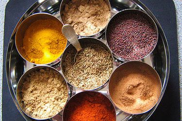 A collection of seven dry spices in a masala dabba, a large tin with smaller tins inside.