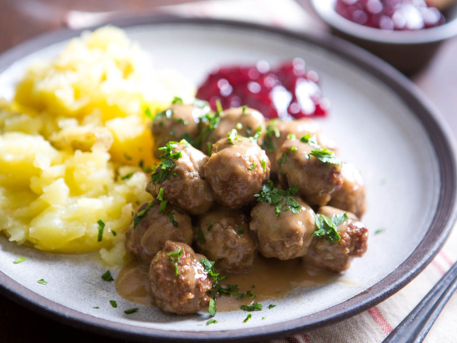 Plate of gravy-coated Swedish meatballs garnished with chopped parsley, boiled potatoes, and lingonberry jam