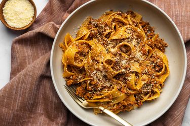 A bowl of pasta with Bolognese sauce on a textured cloth with a small bowl of grated cheese off to the left side.