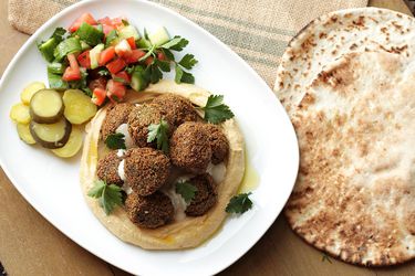 A platter of falafel garnished with flat-leaf parsley, stacked on top of hummus with pickles and cucumber-tomato salad.