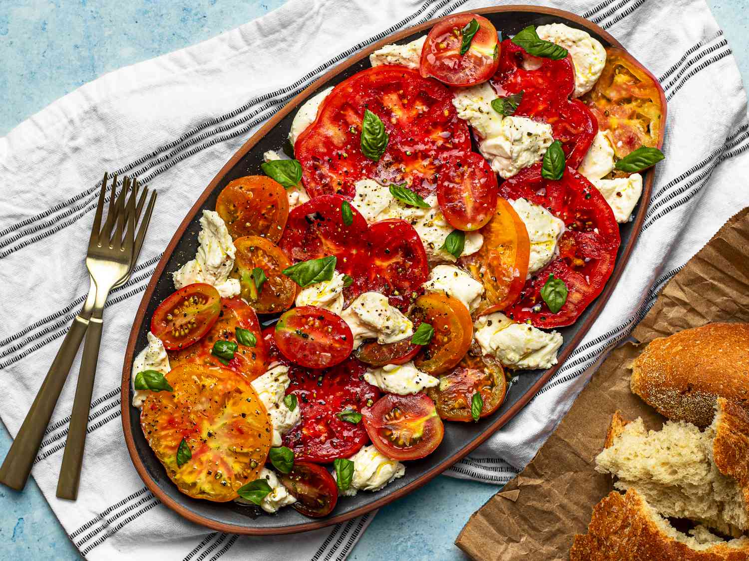 一个n oval ceramic platter holding caprese salad. The platter is on a dish cloth. There is a pair of forks to the left of the platter, and in the bottom right of the image is a wooden cutting board holding torn chunks of bread.