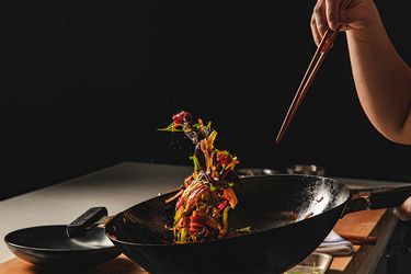 在黑暗的厨房里，一个人拿着筷子扔锅，导致五颜六色的食物悬浮在空气中