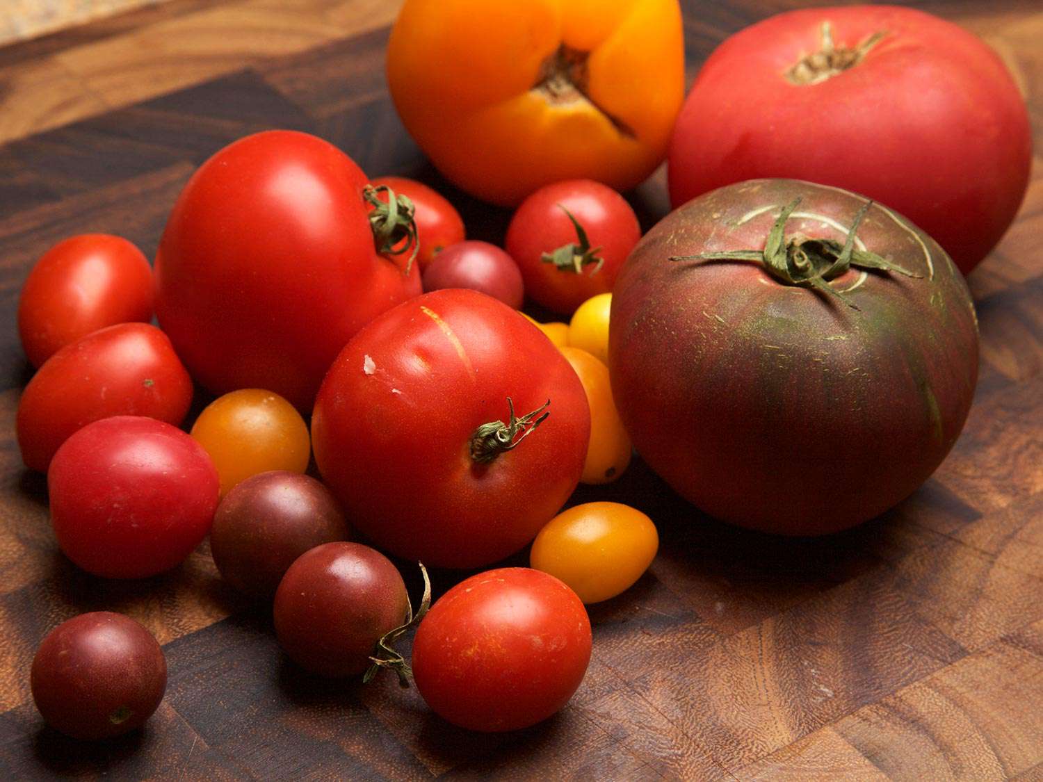 An assortment of ripe tomatoes of different colors and sizes, on a wooden chopping board
