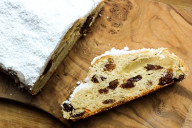 Buttery marzipan stollen dusted with powdered sugar on a wood cutting board. A slice of stollen is laying on the cutting board, exposing the fruit and marzipan inside.