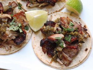 Overhead photograph showing sous vide pork carnitas on corn tortillas topped with salsa verde and cilantro, with lime wedges on the side.