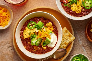 A white speckled ceramic bowl holding chili topped with corn chips, grated cheddar, sour cream, sliced jalapenos, avocado, and scallions. There are saltine crakers on the plate underneath the bowl of chili, and the periphery of the image has small bowls of additional toppings.