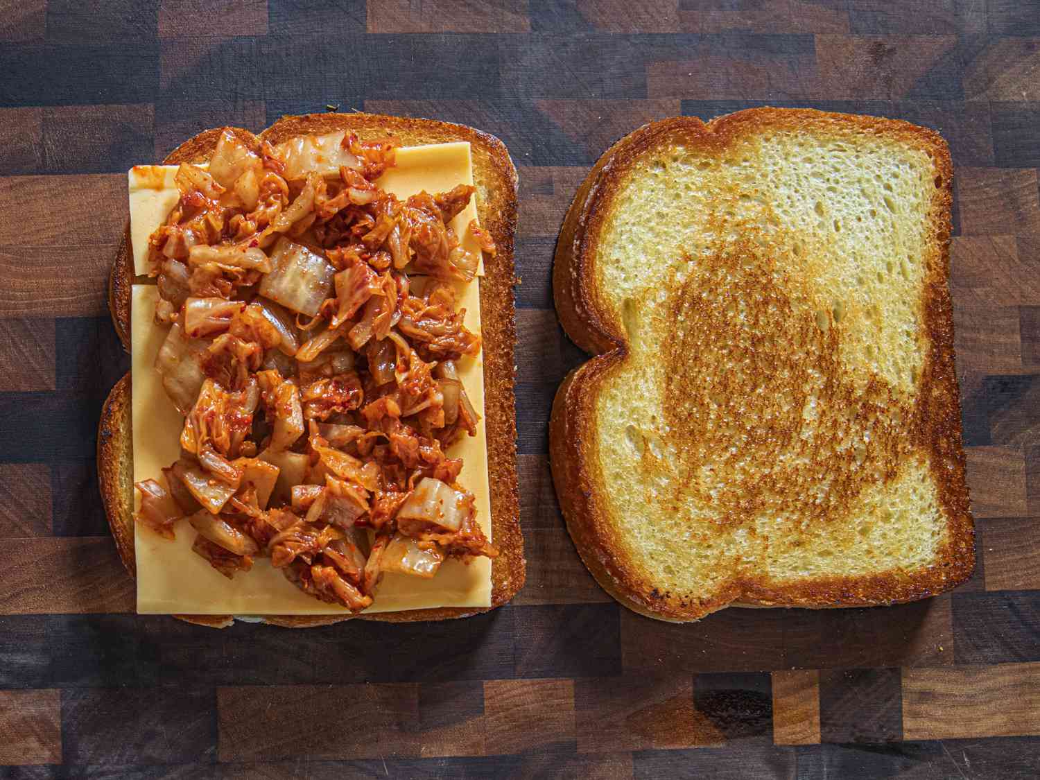 Two pieces of toasted bread on a cutting board with one piece layered with a slice of American Cheese and kimchi