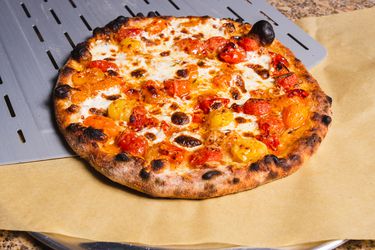 Burst cherry tomato outdoor oven pizza being slid off a peel onto parchment on a pizza pan