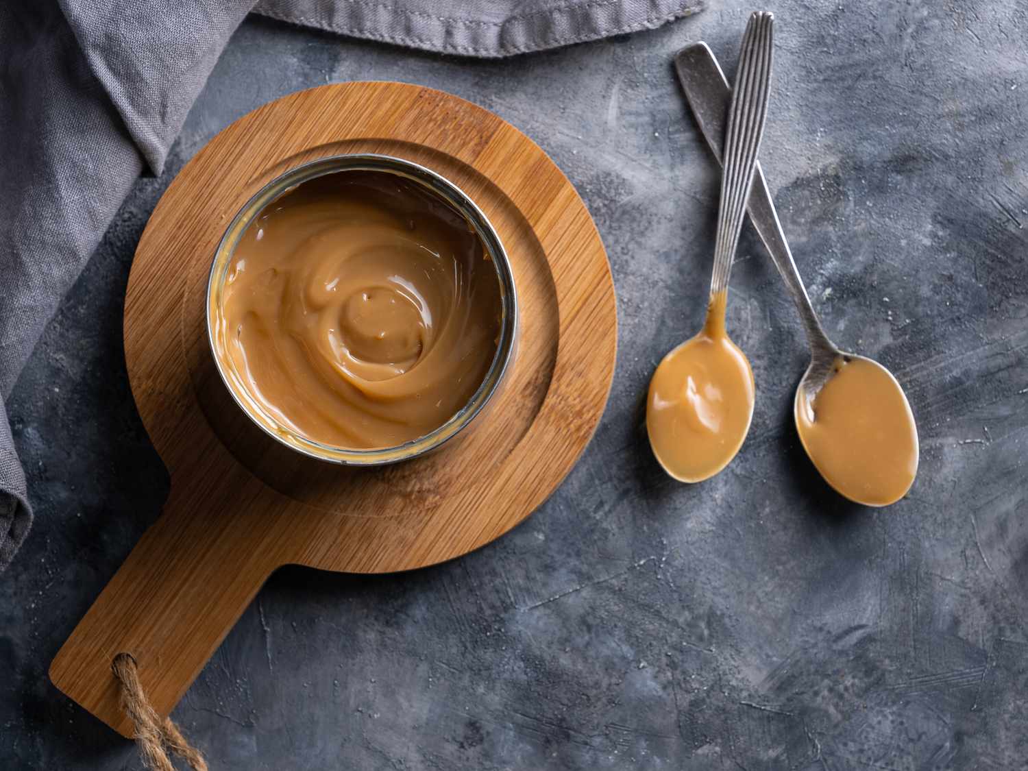 A can of dulce de leche on a wooden trivet with two metal spoons with dulce de leche on them, on a stone background