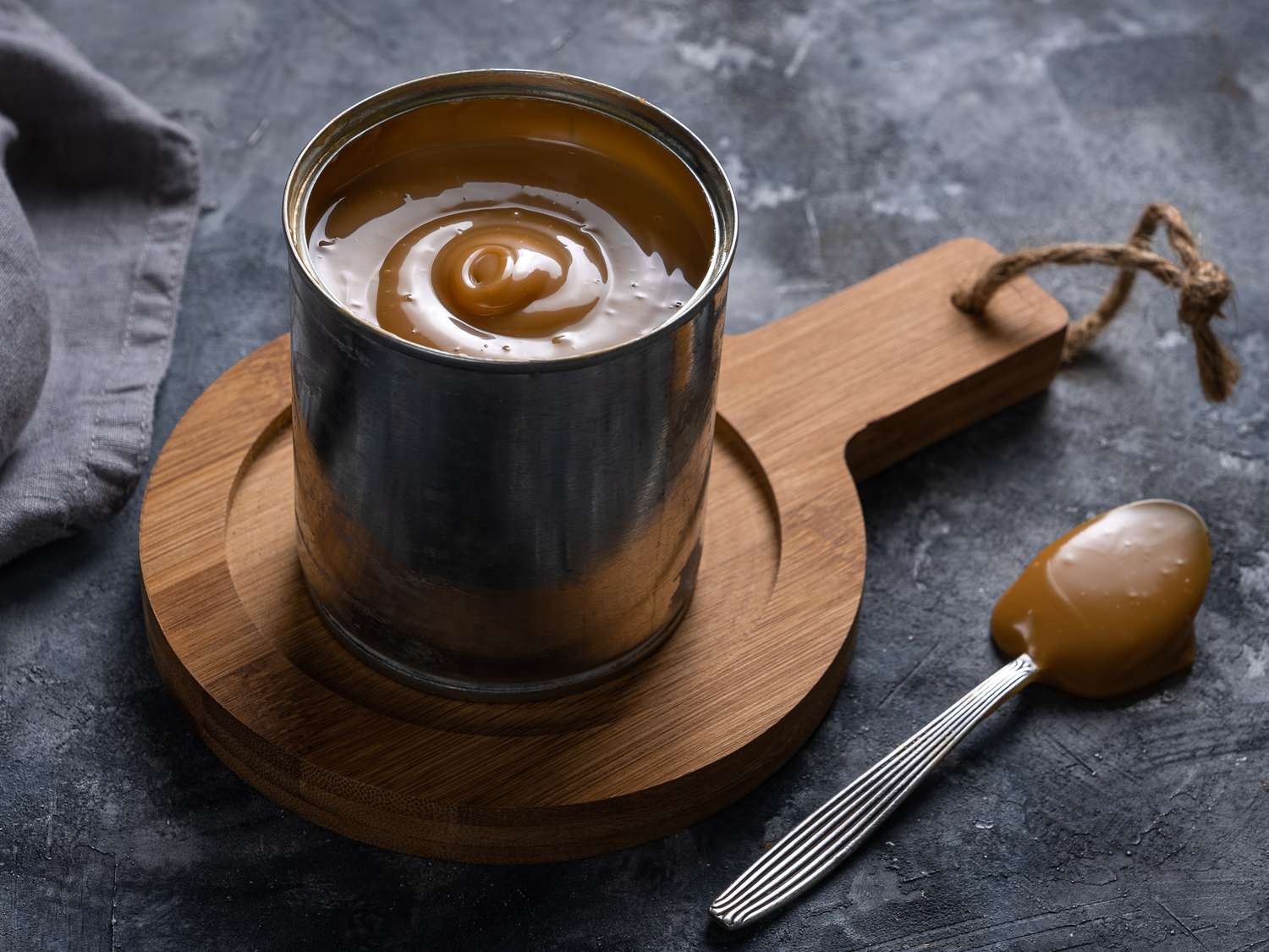 The can of opened dulce de leche on a wooden trivet with a spoon