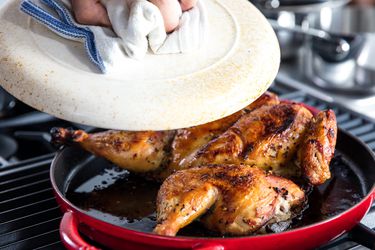 Removing a heavy lid from a golden-brown, crispy whole chicken in a skillet.