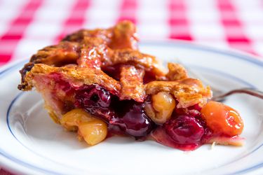 A slice of cherry pie on a white plate with blue edges, sitting on a white and red checkered tablecloth.