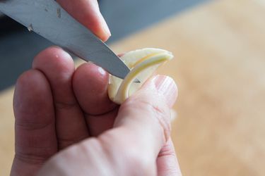 Hands holding a pairing knife and a halved garlic clove. The tip of the pairing knife is prying the germ from the clove.