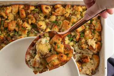 A large copper-colored serving spoon lifting a spoonful of cooked stuffing out of a pan, which is in the background.