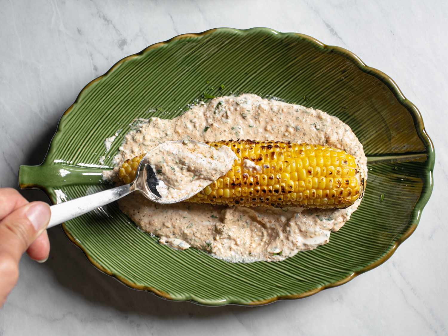 A grilled ear of corn being smothered in sauce for elotes