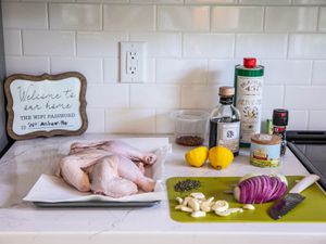 A spread of ingredients on a marble countertop in an Airbnb kitchen: chicken parts on a paper towel–lined tray; onion, garlic, and herbs on a cutting board; balsamic vinegar; fleur de sel; olive oil; grinder of black pepper; lemon halves