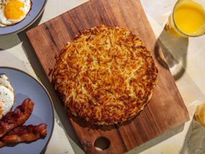 A deeply browned rÃ¶sti sits on a wooden cutting board, with plates of fried eggs and bacon, and glasses of orange juice around it on the table.