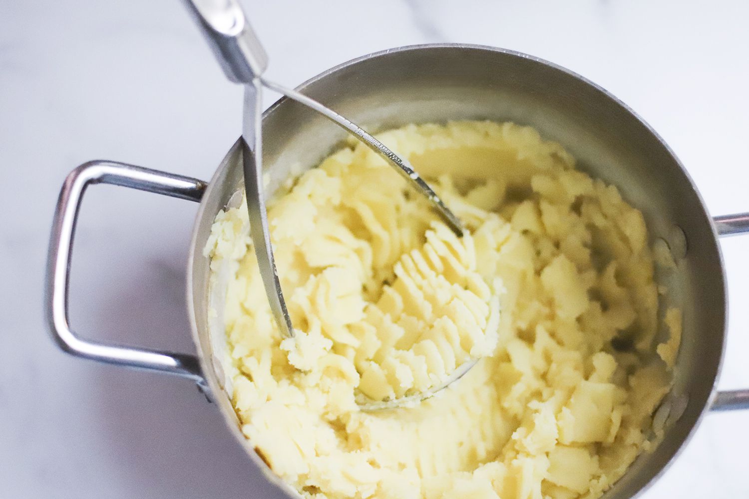 A saucepan with mashed potatoes and a potato masher in it.