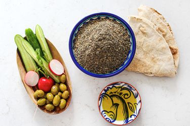 Small bowl of Za'tar next to a piece of pita, a small bowl of olive oil, and a rectangular bowl of vegetables