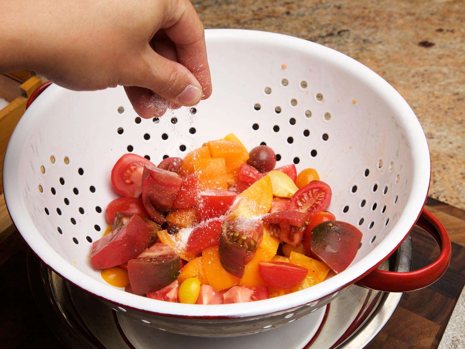 A hand sprinkling salt over chunks of yellow and red ripe tomato, in a colander placed over a bowl