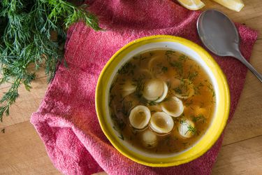 Overhead view of ultimate chicken soup served in a white bowl with bright yellow trim. Fresh dill and lemon wedges are nearby.