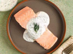 Poached salmon topped with dill-yogurt sauce on a dark plate with a red rim. There is a fork sitting on a napkin in the bottom right corner, and a small bowl of additional sauce in the top left corner.