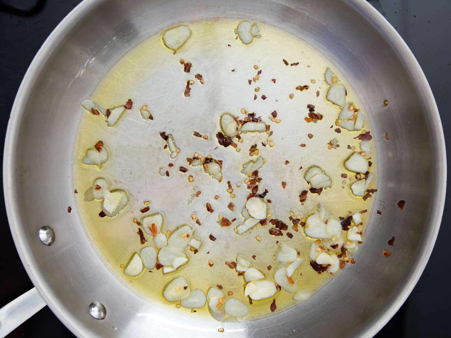 Thinly sliced garlic and red pepper flakes with olive oil inside of a stainless steel skillet.